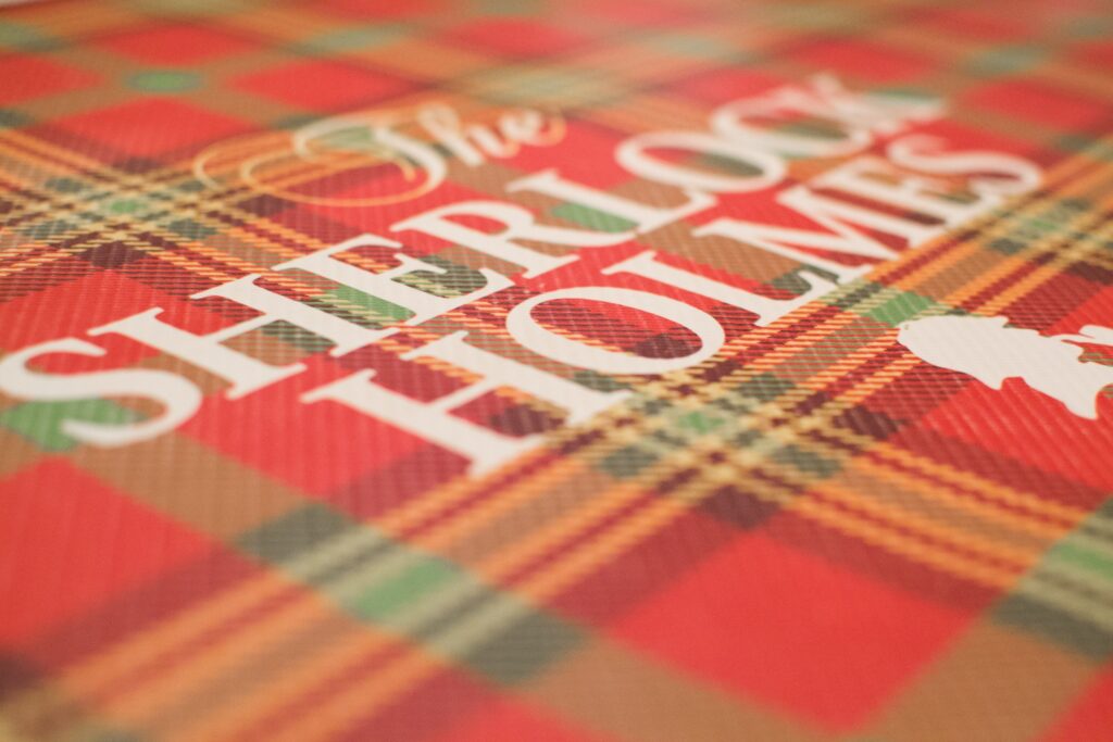 Diseño tartán rojo y verde con el título The Sherlock Holmes en letras blancas y la silueta del detective.
